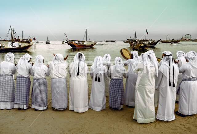 مجموعة من الرجال العرب الخليجيين القطريين على شاطئ بحر الدوحة بقطر ، استقبال البحارة العائدين من رحلة الصيد بالاغاني والدفوف ، وسائل النقل البحرية ، بحر قطر ، قوارب النقل ، سفن خشبية في البحر