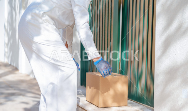 A young Saudi Arabian Gulf man wearing medical gloves to prevent corona virus, delivering orders to customers to the desired place, safe and reliable delivery process, ordering products online, high-quality shipping and delivery services