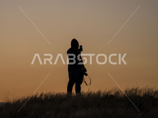 صورة ظلية لمصور بيده كاميرا التصوير ويستخدم الهاتف المحمول، التصوير من خلال الهاتف المحمول، تقنيات حديثة، مناظر طبيعية، النباتات العشبية، تصوير الطبيعة وقت غروب الشمس