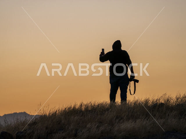 صورة ظلية لمصور بيده كاميرا التصوير ويستخدم الهاتف المحمول، التصوير من خلال الهاتف المحمول، تقنيات حديثة، مناظر طبيعية، النباتات العشبية، تصوير الطبيعة وقت غروب الشمس