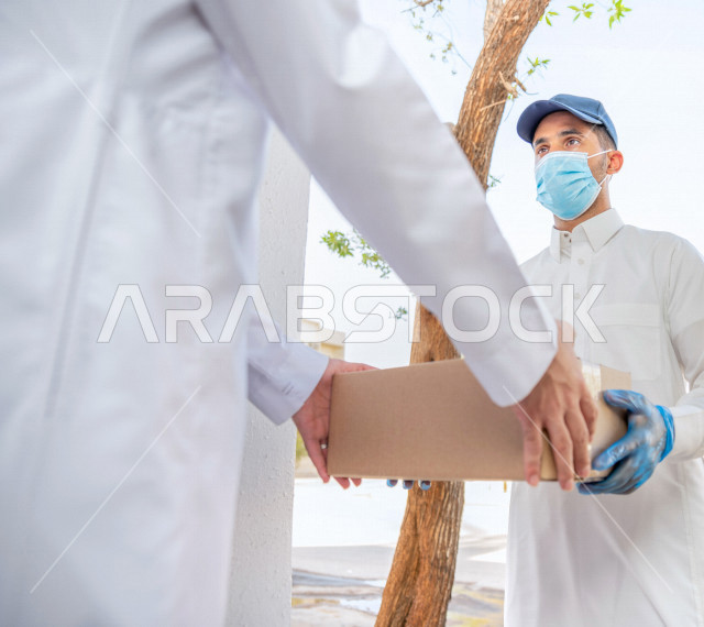شاب عربي سعودي خليجي يرتدي الكمامة والقفازات الطبية للوقاية من فايروس كورونا ، يقوم بتسليم الطلبات للزبائن ، عملية التوصيل الامنة والموثوقة ، طلب المنتجات عبر الانترنت ، خدمات الشحن و التوصيل ذات الجودة العالية