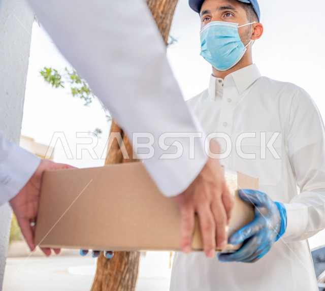 شاب عربي سعودي خليجي يرتدي الكمامة والقفازات الطبية للوقاية من فايروس كورونا ، يقوم بتسليم الطلبات للزبائن ، عملية التوصيل الامنة والموثوقة ، طلب المنتجات عبر الانترنت ، خدمات الشحن و التوصيل ذات الجودة العالية