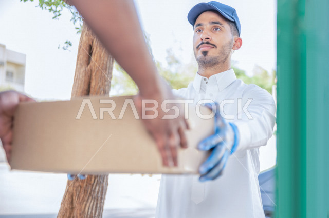 شاب عربي سعودي خليجي يرتدي القفازات الطبية للوقاية من فايروس كورونا ، يقوم بتسليم الطلبات للزبائن ،  عملية التوصيل الامنة والموثوقة ، طلب المنتجات عبر الانترنت ،  خدمات الشحن و التوصيل ذات الجودة العالية