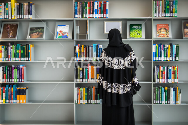 صورة من الخلف لسيدة عربية خليجية قطرية تقف امام رفوف خشبية مليئة بالكتب داخل مكتبة في مدينة الدوحة في قطر، معالم قطر، التعلم والمطالعة، القراءة والتثقف