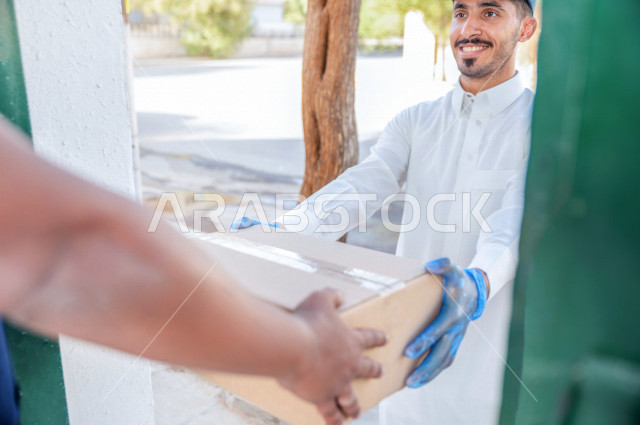 شاب عربي سعودي خليجي يرتدي القفازات الطبية للوقاية من فايروس كورونا ، يقوم بتسليم الطلبات للزبائن ،  عملية التوصيل الامنة والموثوقة ، طلب المنتجات عبر الانترنت ،  خدمات الشحن و التوصيل ذات الجودة العالية