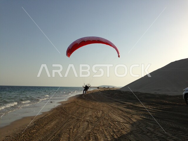 رجل عربي خليجي قطري يمارس رياضة الطيران الشراعي، شراع بالوان علم قطر، تل رملي، شاطئ بحر خور العديد، انشطة ترفيهية، السياحة في قطر
