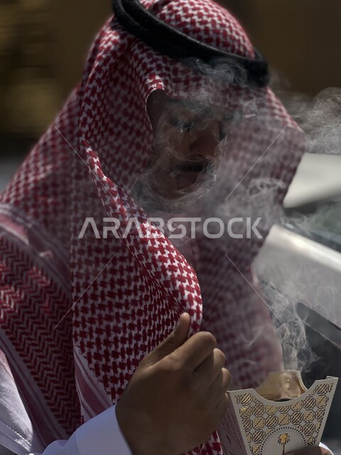 صورة مقربة لرجل عربي سعودي  يحمل حامل البخور( المبخرة ) ، استخدام خشب العود في المناسبات الاجتماعية و المناسبات الدينية ،  اصالة العادات و التقاليد في المملكة العربية السعودية ، التطيب و التعطر .