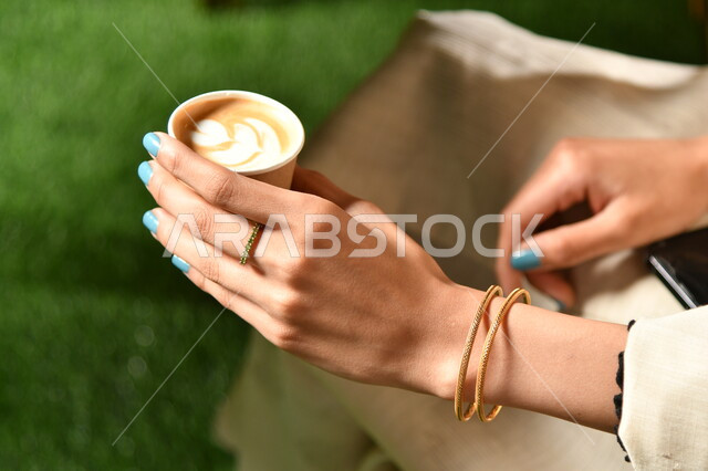 A picture from above of a Saudi Arab woman's hand holding a cup of coffee with thick foam with graphics, hot cappuccino drink, delicious hot drinks, hot coffee cup, grass background.