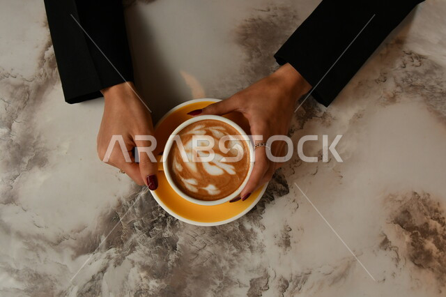 A picture from above of a woman's hands holding a cup of coffee with thick foam, hot cappuccino drink, delicious hot drinks, hot coffee cup, gray background.