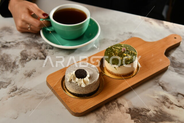 يد امرأة عربية سعودية  تمسك بفنجان القهوة الداكنة, مشروبات ساخنة لذيذة, فنجان القهوة الساخن, قطع التشيز كيك المزينة على لوح خشبي .