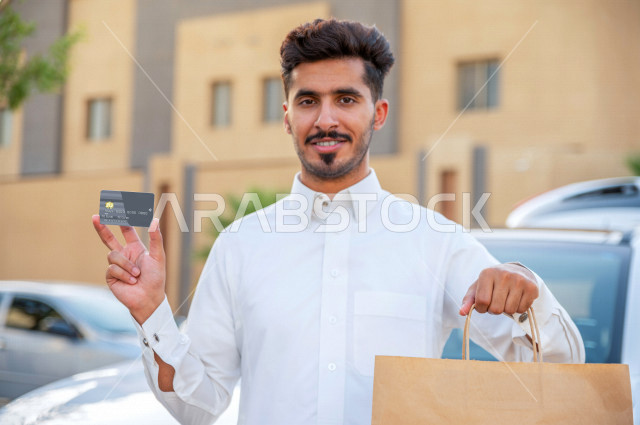 A smiling Saudi Arabian Gulf young man holding a delivery parcel and credit card in his hand, credit card payment upon receipt, the process of delivering orders to customers, high-quality shipping and delivery services