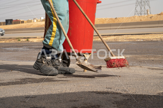 صورة عامل النظافة باللباس الخاص بالعمل يقوم بإزالة النفايات من الطرقات و الأرصفة ، عربة التنظيف ، أدوات النظافة الخاصة بالعمل ، الاهتمام بنظافة البيئة، بلدية مدينة الرياض.