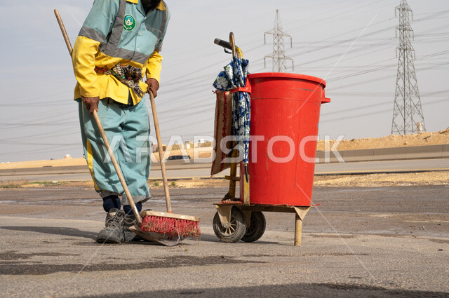 Image of a cleaning worker in work clothes cleaning roads and sidewalks, cleaning cart and cleaning tools for work, caring for environmental cleanliness, Riyadh Municipality.