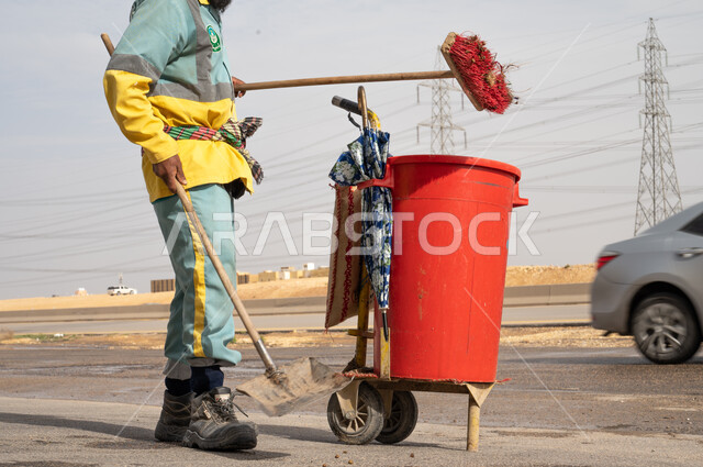 صورة عامل النظافة بالزي الخاص بالعمل يقوم إزالة النفايات والقمامة من الطريق ، عربة التنظيف  وأدوات النظافة الخاصة بالعمل ، الاهتمام بنظافة البيئة ، بلدية مدينة الرياض.
