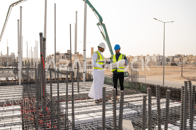 مهندسان سعوديان عاملان يقفان داخل موقع البناء على قواطع الاسمنت و يحملان مخطط لمشروع البناء في المملكة العربية السعودية , إرتداء خوذة وسترة الحماية الخاصة بالعمل, تدقيق العمل .