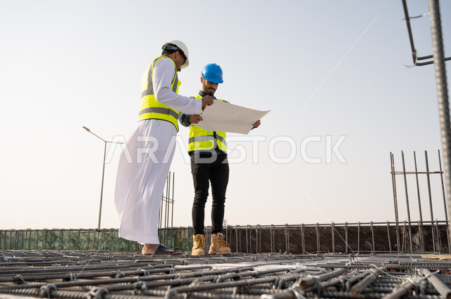 صورة من الاسفل لمهندسان سعوديان عاملان يحملان مخطط لمشروع البناء للتخطيط والتنفيذ  في المملكة العربية السعودية , إرتداء خوذة وسترة الحماية الخاصة بالعمل ,  خوذة السلامة الزرقاء, المهندس المشرف بخوذة بيضاء , تدقيق العمل .