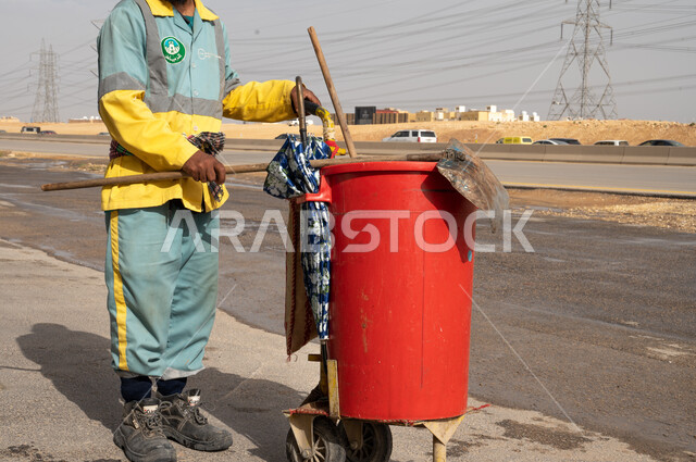صورة مقربة لعامل النظافة باللباس الخاص بالعمل ، تنظيف  الطرقات  والاهتمام بنظافة البيئة ، عربة التنظيف  وأدوات النظافة الخاصة بالعمل ، بلدية مدينة الرياض.