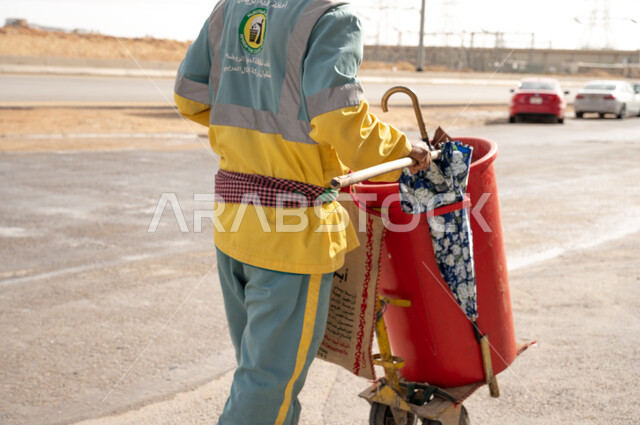 عامل النظافة بصورة مقربة  بالزي الخاص بالعمل ، تنظيف  الطرقات والأرصفة  والاهتمام بنظافة البيئة ، عربة التنظيف  وأدوات النظافة الخاصة بالعمل ، بلدية مدينة الرياض.