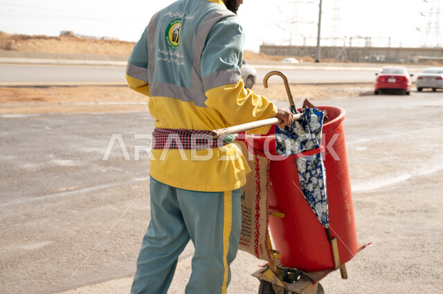 عامل النظافة بصورة مقربة  بالزي الخاص بالعمل ، إزالة النفايات والقمامة من الطرقات والأرصفة  والاهتمام بنظافة البيئة ، عربة التنظيف وأدوات النظافة الخاصة بالعمل ، بلدية مدينة الرياض.