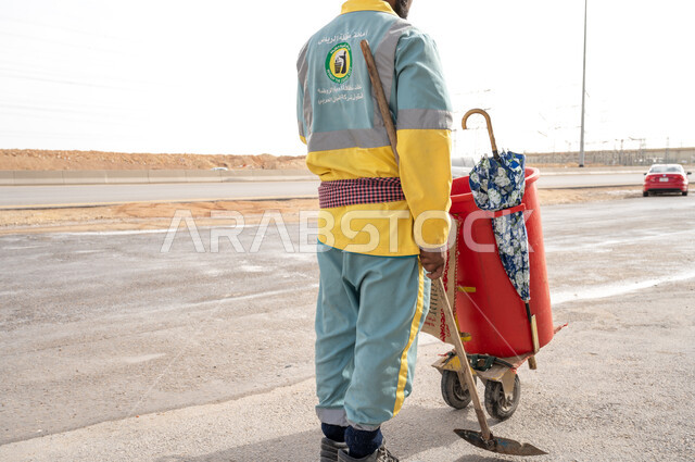 عامل النظافة  بالزي الخاص بالعمل بصورة مقربة  ، تنظيف الطرقات والأرصفة  والاهتمام بنظافة البيئة ، عربة التنظيف وأدوات النظافة الخاصة بالعمل ، بلدية مدينة الرياض.