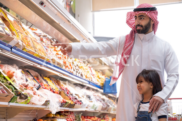  رجل عربي سعودي خليجي في متجر الفواكه برفقة طفلته داخل السوبرماركت ، الاستمتاع بالشراء والتسوق ، السلع والمواد الغذائية ، مستلزمات المستهلك.