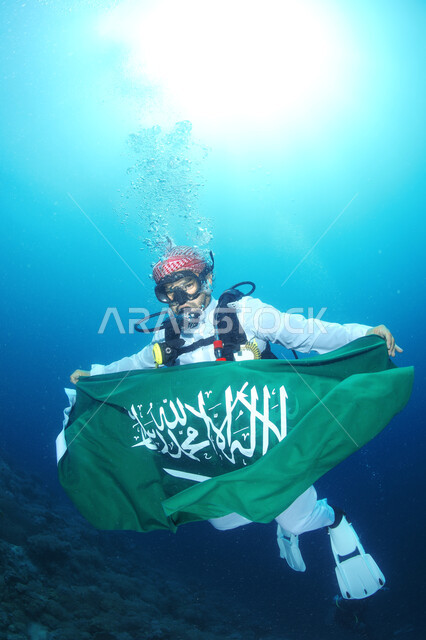 A Saudi Arabian Gulf diver wearing the traditional Saudi dress and underwater diving suit, holding the flag of the Kingdom of Saudi Arabia, the 92nd anniversary of the Saudi National Day
