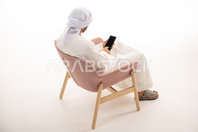 Portrait from the back of an Arab Gulf Emirati man sitting on a chair, using the phone, social communication, conversations and chatting, communicating with friends, a remote work meeting, a white background