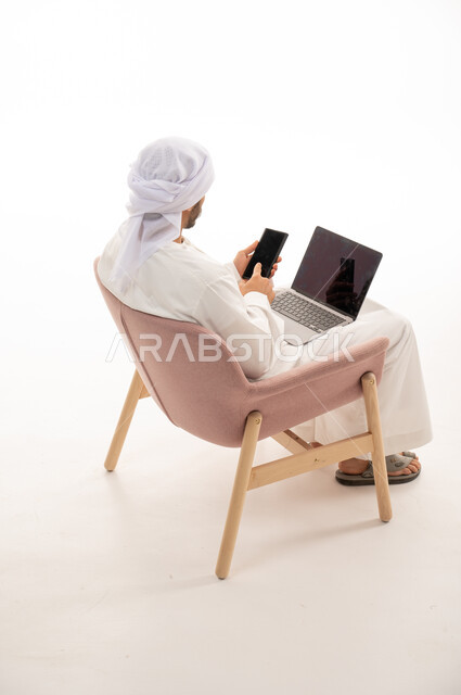 بورتريه من الخلف لرجل عربي خليجي إماراتي يجلس على كرسي يستخدم جهاز الحاسوب والهاتف المحمول، اجراء مكالمات فيديو ، محادثات ودردشة، تواصل اجتماعي،  التواصل مع الأصدقاء، اجتماع عمل عن بعد،  خلفية بيضاء .