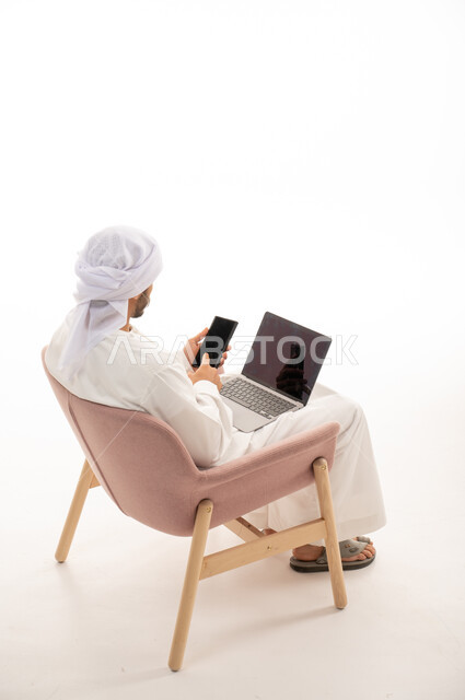 بورتريه من الخلف لرجل عربي خليجي اماراتي يجلس على كرسي يستخدم جهاز الحاسوب والهاتف المحمول، اجراء مكالمات فيديو، تواصل اجتماعي، محادثات ودردشة، التواصل مع الأصدقاء، اجتماع عمل عن بعد،  خلفية بيضاء .