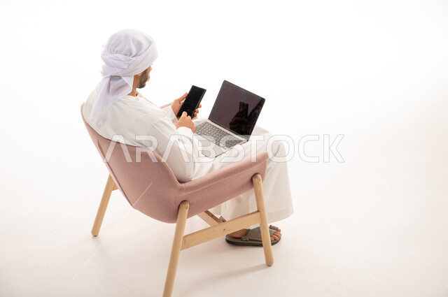 بورتريه من الخلف لرجل عربي خليجي إماراتي يجلس على كرسي يستخدم جهاز الحاسوب والهاتف المحمول، تواصل اجتماعي،  اجراء مكالمات فيديو ، محادثات ودردشة، التواصل مع الأصدقاء، اجتماع عمل عن بعد،  خلفية بيضاء .