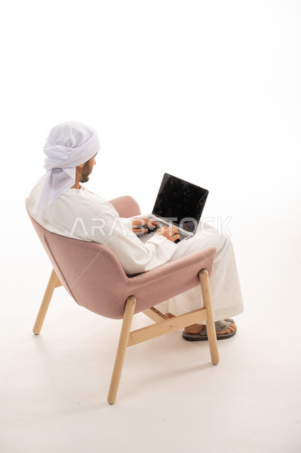 بورتريه من الخلف لرجل عربي خليجي إماراتي يجلس على كرسي يستخدم جهاز الحاسوب، اجراء مكالمات فيديو، تواصل اجتماعي، محادثات ودردشة، التواصل مع الأصدقاء، تصفح الانترنت , اجتماع عمل عن بعد،  خلفية بيضاء .