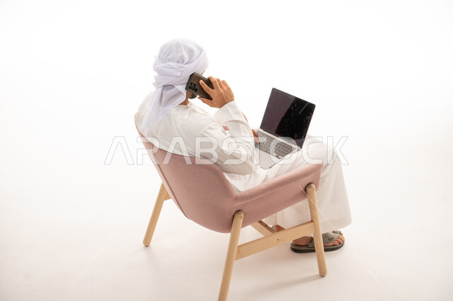بورتريه من الخلف لرجل عربي خليجي إماراتي يجلس على كرسي يستخدم جهاز الحاسوب، اجراء مكالمات هاتفية ، تواصل اجتماعي، محادثات ودردشة، التواصل مع الأصدقاء، اجتماع عمل عن بعد ،  خلفية بيضاء .