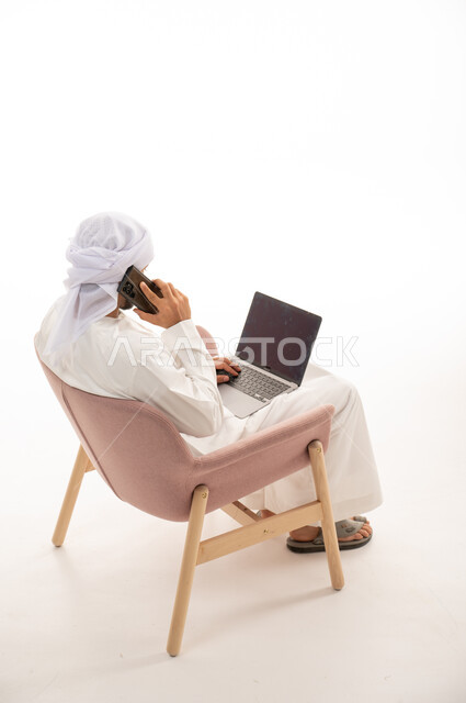 بورتريه من الخلف لرجل عربي خليجي إماراتي يجلس على كرسي يستخدم جهاز الحاسوب، إجراء مكالمات هاتفيه ، تواصل اجتماعي، محادثات ودردشة، التواصل مع الأصدقاء، اجتماع عمل عن بعد ،  خلفية بيضاء .