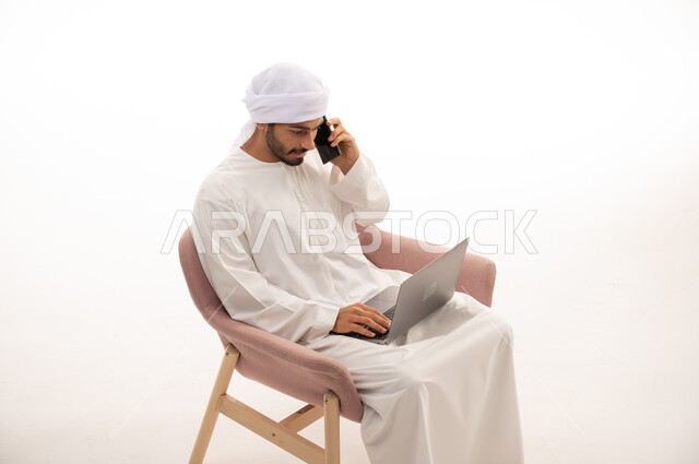 بورتريه لرجل عربي خليجي إماراتي يجلس على كرسي يستخدم جهاز الحاسوب، اجراء مكالمات هاتفيه ، تواصل اجتماعي، محادثات ودردشه ، التواصل مع الأصدقاء، اجتماع عمل عن بعد ،  خلفية بيضاء .