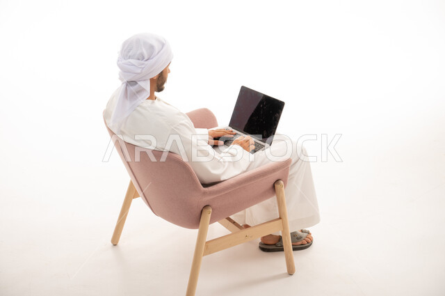 بورتريه من الخلف لرجل عربي خليجي إماراتي يجلس على كرسي يستخدم جهاز الحاسوب، اجراء مكالمات فيديو، تواصل اجتماعي، محادثات ودردشة ، اجتماع عمل عن بعد ،التواصل مع الأصدقاء،  تصفح الانترنت , خلفية بيضاء .