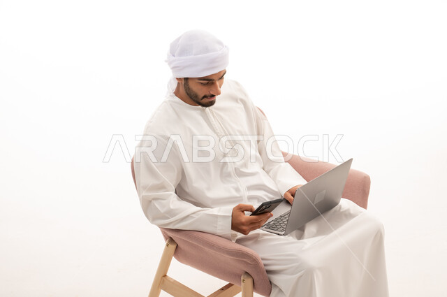 بورتريه  لرجل عربي خليجي إماراتي يجلس على كرسي يستخدم جهاز الحاسوب والهاتف المحمول، تواصل اجتماعي، محادثات ودردشة، التواصل مع الأصدقاء، اجراء مكالمات فيديو، اجتماع عمل عن بعد،  خلفية بيضاء .