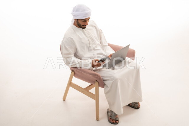 بورتريه  لرجل عربي خليجي إماراتي يجلس على كرسي , استخدام جهاز الحاسوب والهاتف المحمول، اجراء مكالمات فيديو، تواصل اجتماعي، محادثات ودردشة، التواصل مع الأصدقاء، اجتماع عمل عن بعد،  خلفية بيضاء .