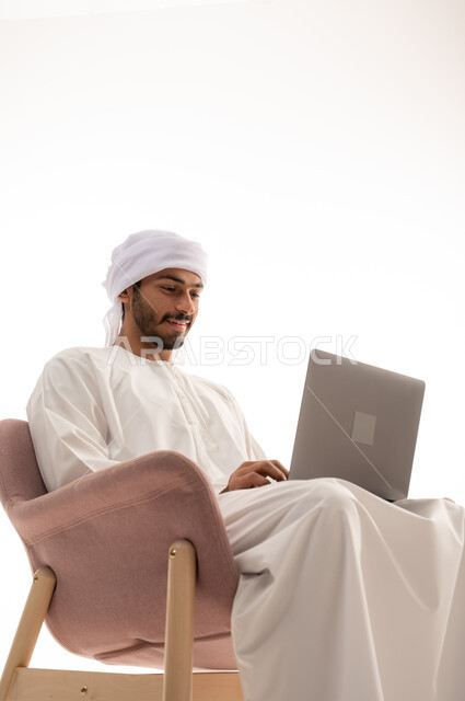 بورتريه لرجل عربي خليجي اماراتي يجلس على كرسي يستخدم جهاز الحاسوب، اجراء مكالمات فيديو، تواصل اجتماعي، محادثات ودردشة، التواصل مع الأصدقاء، اجتماع عمل عن بعد،  تصفح الانترنت ، خلفية بيضاء .