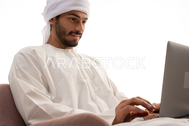 Portrait of an Arab Gulf Emirati man sitting on a chair using a computer, making video calls, social communication, conversations and chatting, communicating with friends, a remote business meeting, surfing the Internet, a white background
