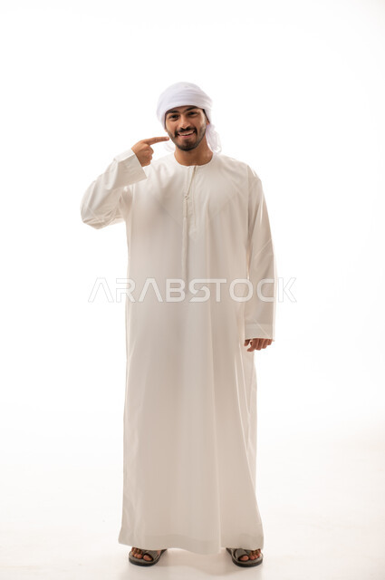 بورتريه لرجل عربي اماراتي خليجي يبتسم ويشير بإصبعه السبابة على أسنانه،  إبتسامة هوليود، استخدام قشور فينير من الخزف للأسنان، تجميل وتبييض الاسنان، معالجة الأسنان،  أسنان نظيفة مثالية , صورة الجسم بالطول الكامل , خلفية بيضاء 