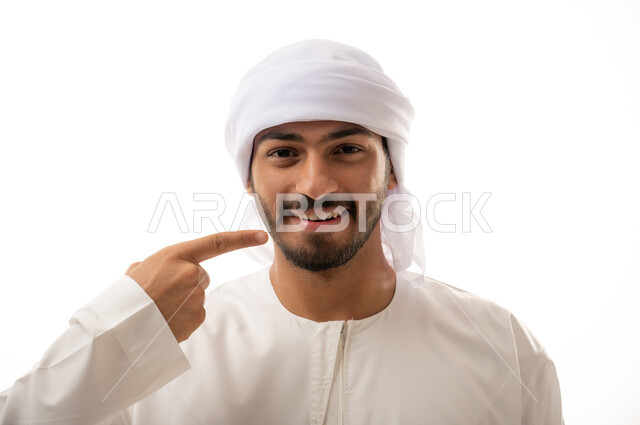 بورتريه لرجل عربي اماراتي خليجي يبتسم ويشير بإصبعه السبابة على أسنانه،  استخدام قشور فينير من الخزف للأسنان، إبتسامة هوليود، تجميل وتبييض الاسنان، معالجة الأسنان،  أسنان نظيفة مثالية ,خلفية بيضاء 