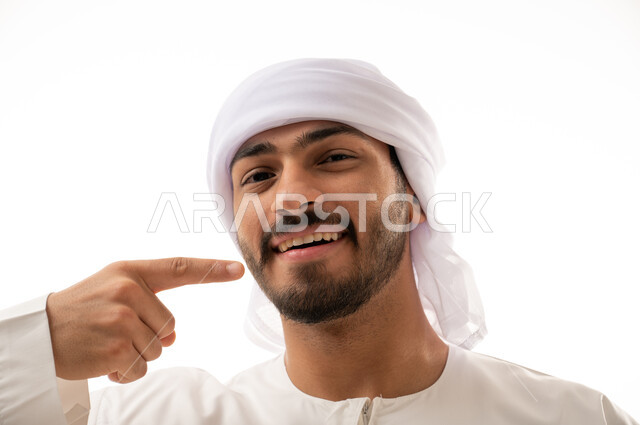 بورتريه لرجل عربي اماراتي خليجي يبتسم ويشير بإصبعه السبابة على أسنانه،  إبتسامة هوليود، استخدام قشور فينير من الخزف للأسنان،  معالجة الأسنان،  أسنان نظيفة مثالية ,تجميل وتبييض الاسنان، خلفية بيضاء 