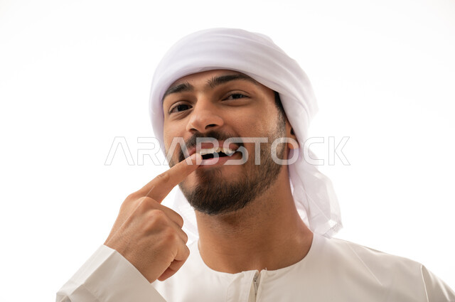 بورتريه لرجل عربي اماراتي خليجي يبتسم ويشير بإصبعه السبابة على أسنانه،  إبتسامة هوليود، تجميل وتبييض الاسنان، معالجة الأسنان،  أسنان نظيفة مثالية , استخدام قشور فينير من الخزف للأسنان،خلفية بيضاء 