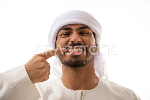 بورتريه لرجل عربي اماراتي خليجي يبتسم ويشير بإصبعه السبابة على أسنانه،  إبتسامة هوليود ، أسنان نظيفة مثالية ,  استخدام قشور فينير من الخزف للأسنان، تجميل وتبييض الاسنان، معالجة الأسنان،  خلفية بيضاء 