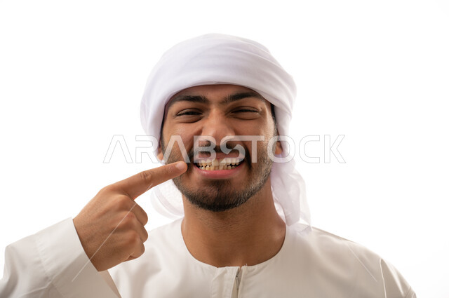 بورتريه لرجل عربي اماراتي خليجي يبتسم ويشير بإصبعه السبابة على أسنانه،  إبتسامة هوليود، استخدام قشور فينير من الخزف للأسنان،   أسنان نظيفة مثالية , تجميل وتبييض الاسنان، معالجة الأسنان، خلفية بيضاء 