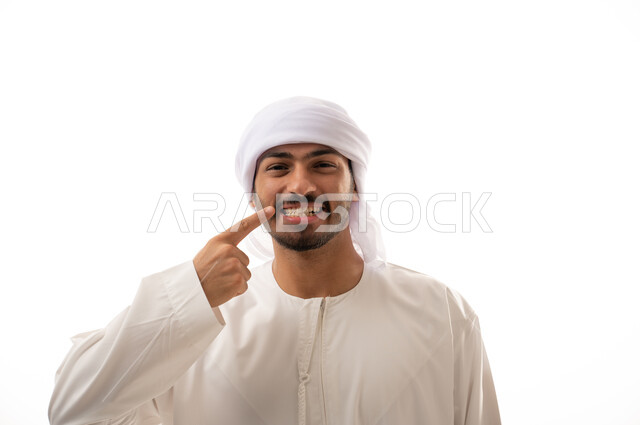 بورتريه لرجل عربي اماراتي خليجي يبتسم ويشير بإصبعه السبابة على أسنانه،  إبتسامة هوليود، استخدام قشور فينير من الخزف للأسنان، تجميل وتبييض الاسنان، معالجة الأسنان،  أسنان نظيفة مثالية ,خلفية بيضاء 