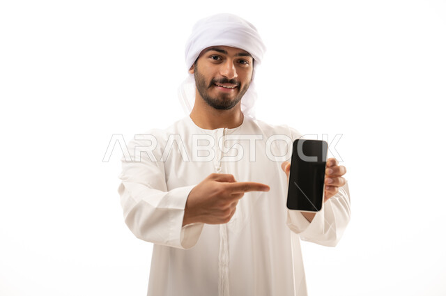 بورتريه لرجل عربي اماراتي خليجي مبتسم ، يشير بإصبعه السبابة على الهاتف المحمول ، عرض شاشة سوداء فارغة ، عرض البرامج والتطبيقات ، التسوق الإلكتروني ، خلفية بيضاء .