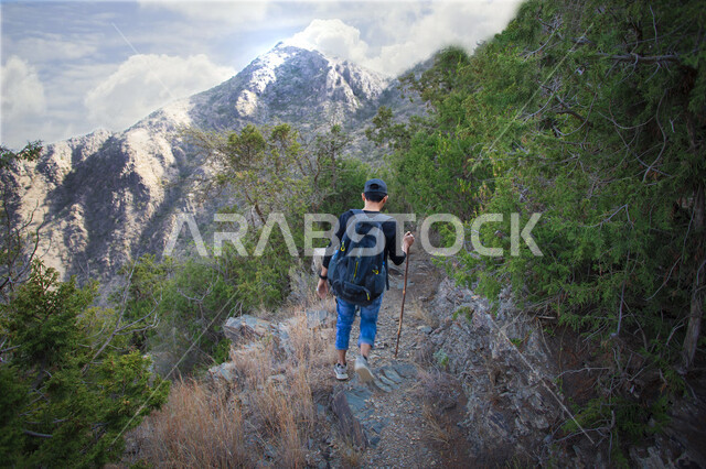 صورة من الخلف لشاب عربي خليجي سعودي يمارس رياضة الهايكنج في جبال عسير في المملكة العربية السعودية، الاشجار والنباتات الخضراء، المشي على الجبال، قمم جبلية، مناظر طبيعية، صحة ولياقة بدنية
