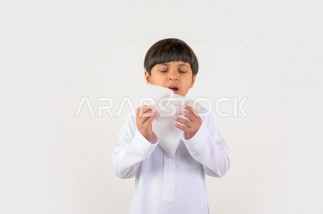 Portrait of a Saudi Arabian Gulf child wearing the Saudi dress, hand and face gestures indicating sneezing, feeling sick, cold, using tissues for sneezing, Corona (Covid_19)