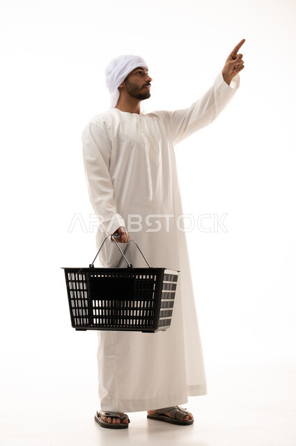 Portrait of a young Emirati from the Gulf, dressed in traditional Emirati clothing, doing shopping, shopping cart, pointing his finger at the top, buying household needs, various goods and foodstuffs, consumer necessities, full-length body image, white ba
